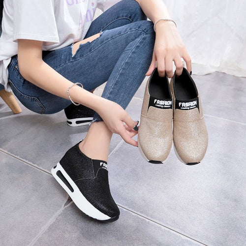 A person holding beige slip-on wedge sneakers with a black elastic band labeled FASHION, wearing black ones.