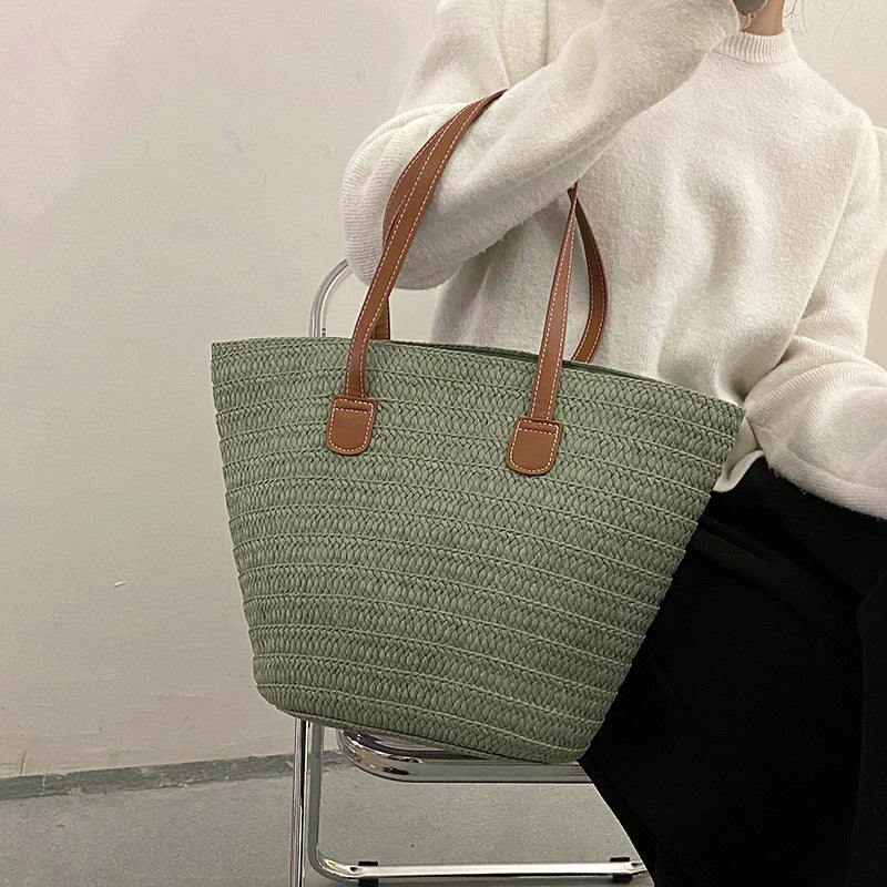 Sage green woven straw tote bag with brown leather handles, held by a person.