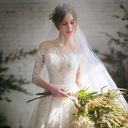 Bride in an off-white lace wedding dress with long sleeves, veil, and flower crown holding a bouquet.