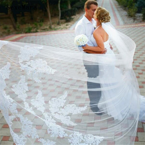 A long white lace-trimmed bridal veil flows dramatically in the foreground of an outdoor wedding photo.