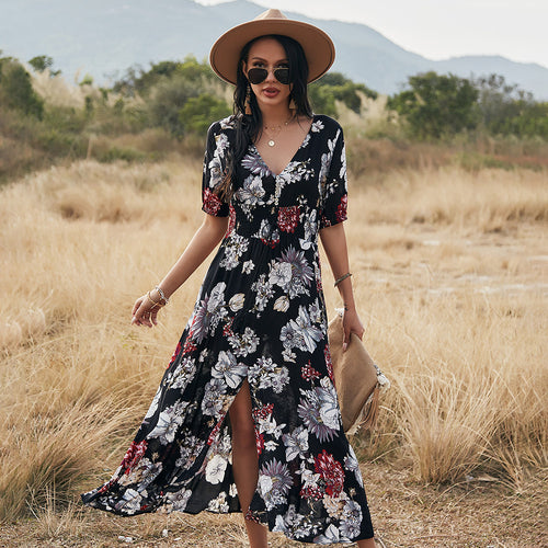 A model in a black V-neck floral dress with short sleeves, a flared skirt, and a front slit, posing in a field.
