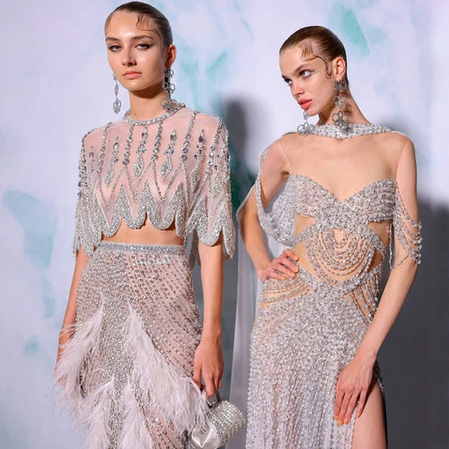 Two models wearing glittering, crystal-embellished evening wear with feathers and sheer fabrics.