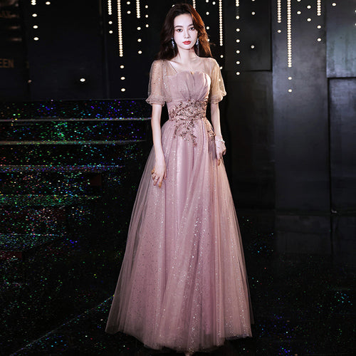 A woman wearing an elegant, floor-length pink evening dress with sheer sleeves, a fitted bodice, and sparkling tulle.