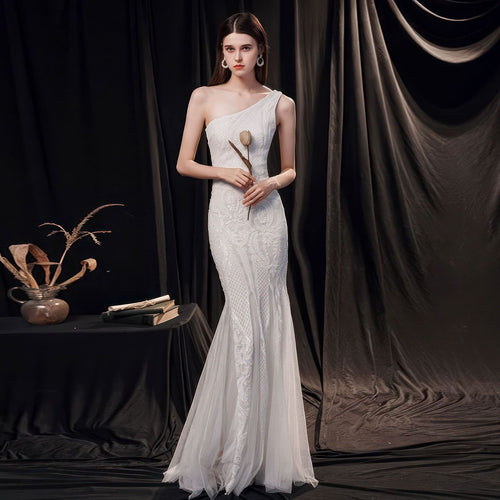 A woman poses in a white, one-shoulder fishtail gown with lace patterns, holding a single dry flower.