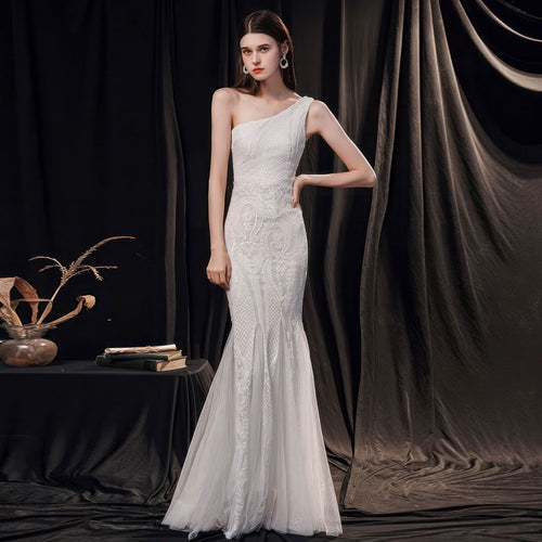 A woman wearing a white, one-shoulder, form-fitting fishtail dress with lace embroidery, standing indoors.