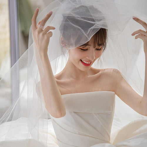 A smiling bride in a white satin strapless gown holding a sheer veil over her head.