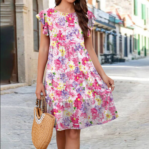 A person models a pink, tiered floral dress with flutter sleeves and holds a straw bag outdoors.